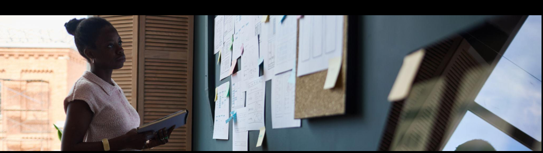 Black woman entrepreneur reviewing notes and planning systems on a wall board while reflecting on accountability, structure, and long-term execution.